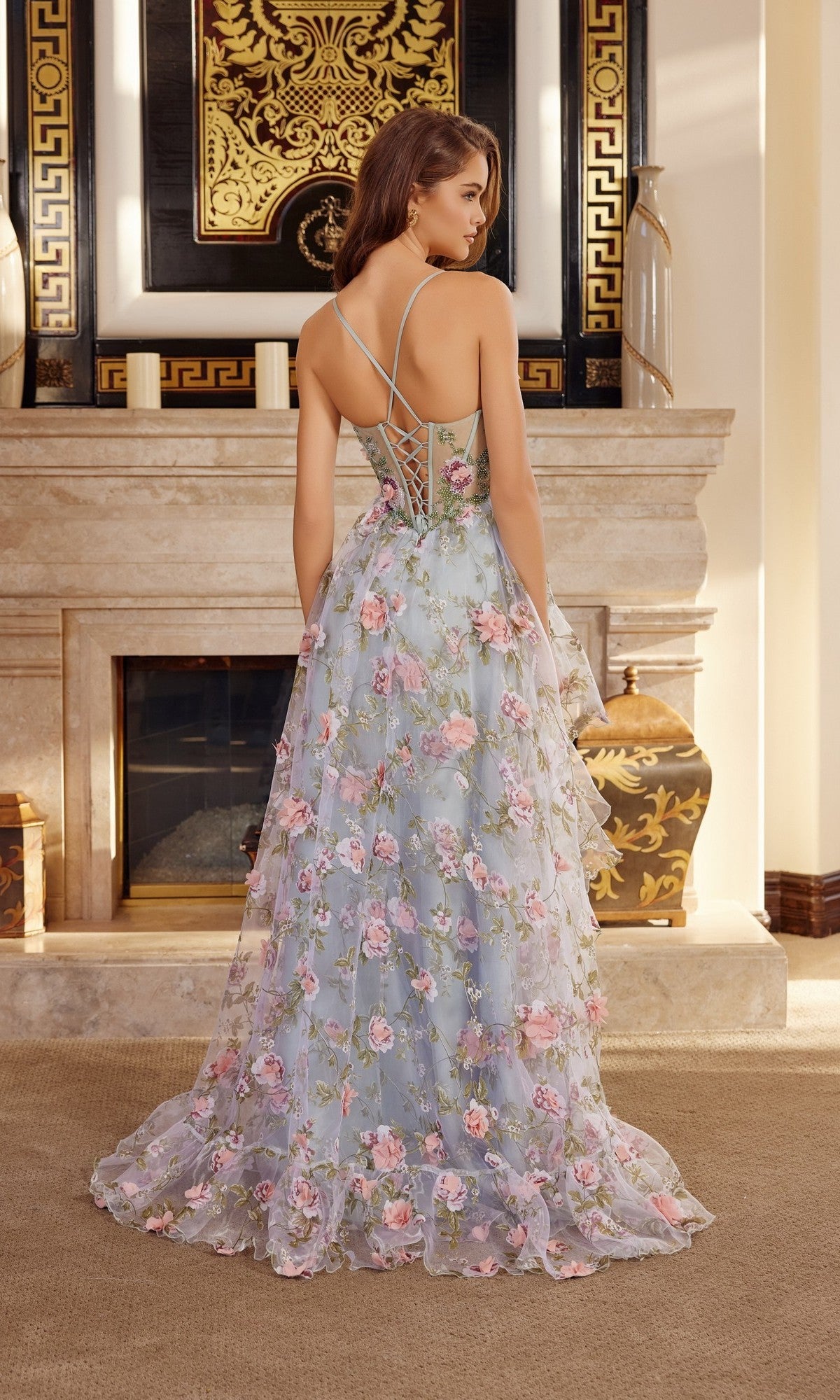 A woman indoors faces away, wearing the Nox Anabel G1783 long prom dress with blue floral chiffon, thin straps, and a sheer back, standing before a marble fireplace and decorative vases.